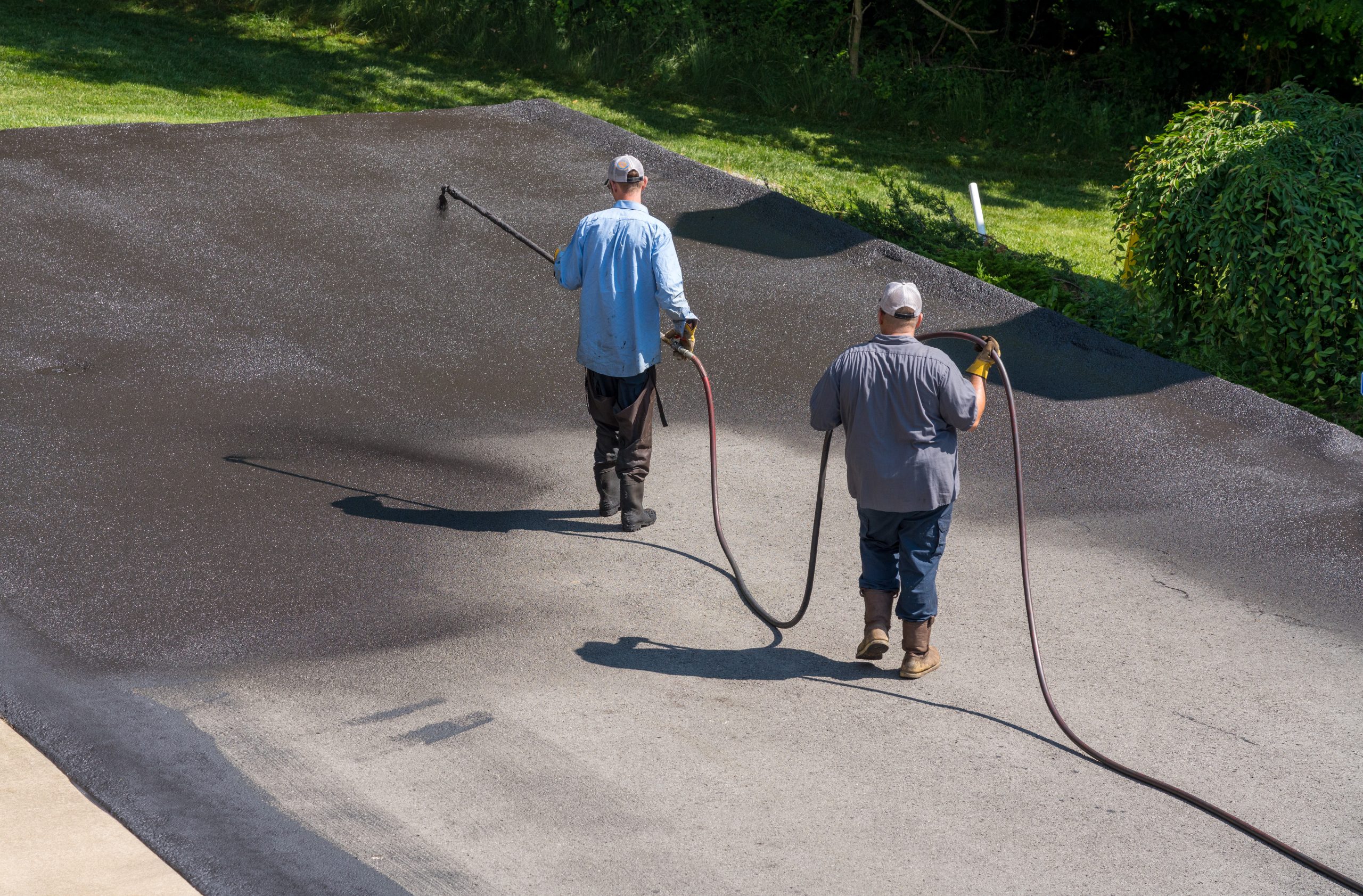 Two workers applying blacktop sealcoat to asphalt driveway using spray equipment for surface protection and durability.