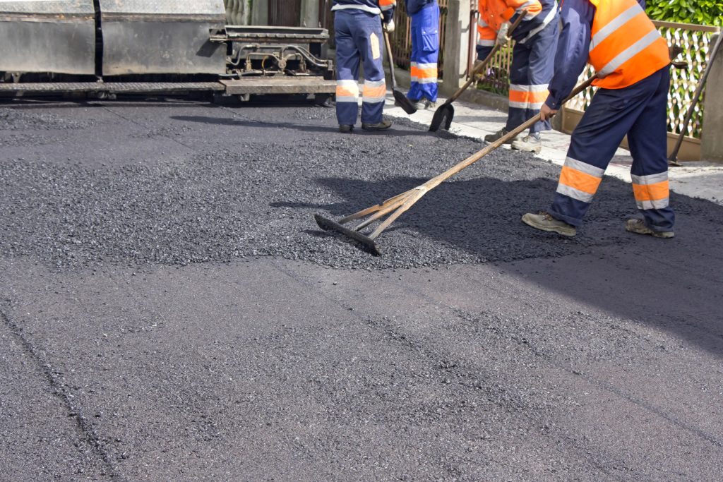Road construction workers spreading hot asphalt during paving with rakes and safety gear.